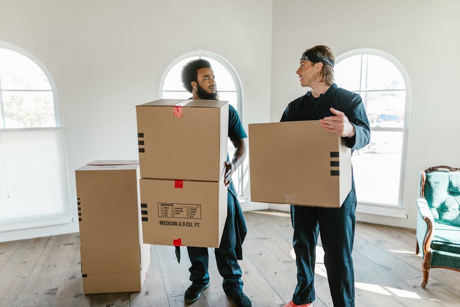 Two professional movers from Man With a Van Clapton are inside a well-lit, spacious room with hardwood flooring and arched windows. One mover, an African-American man, is standing upright and holding two stacked cardboard boxes, each marked with handling labels and sizes, while the other, a Caucasian man with long hair and a headband, is engaged in conversation and carrying a single cardboard box. Additional boxes of various sizes and materials, including some wrapped in plastic and others in plain cardboard, are placed on the floor nearby, indicating packing or packaging for a house removal. The environment appears organized and prepared for a furniture transport or home relocation process, with natural daylight streaming through the windows, highlighting the indoor setting and the preparation work involved in packing and moving services.