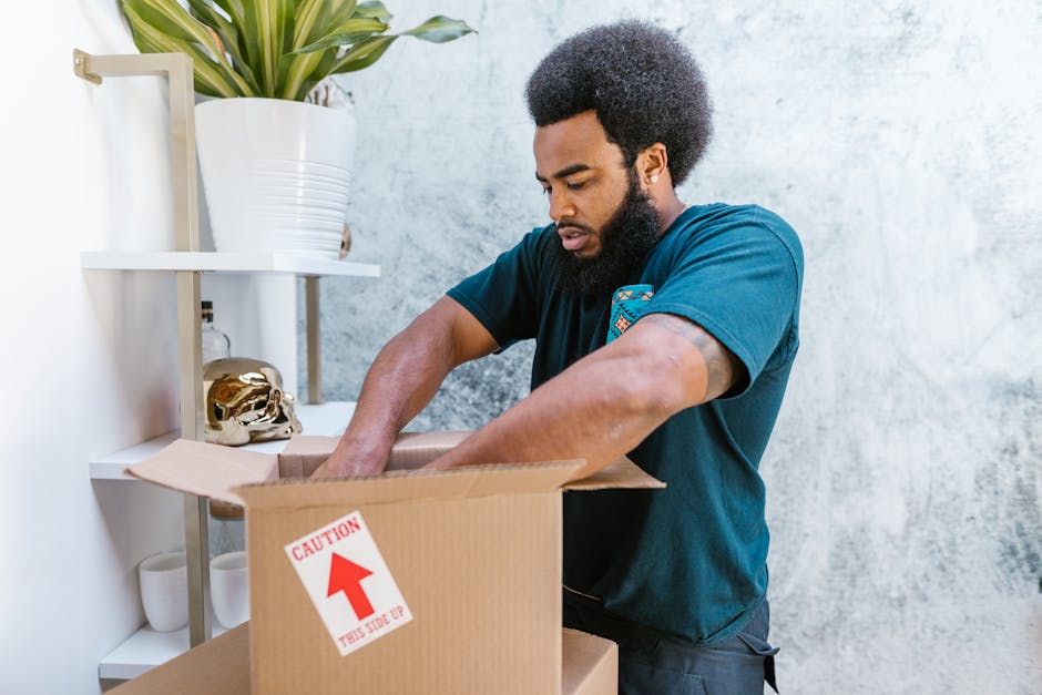 A man with dark skin, an afro hairstyle, and a beard is shown packing a cardboard box with packing materials inside a room. He is wearing a dark blue T-shirt and is reaching into the open box, ensuring items are properly placed for transportation. Behind him, there is a white shelving unit with a large potted plant on the top shelf and decorative objects, including a skull-shaped ornament, on subsequent shelves. The setting appears well-lit with natural light, and the wall behind features a light, textured finish. This scene captures the process involved in home relocation or packing as part of a professional removals service, exemplified by Man With a Van Clapton, supporting efficient and organized furniture transport and packing and moving logistics.