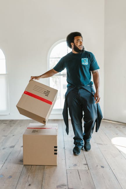 Two professional movers from Man With a Van Clapton are inside a well-lit, spacious room with hardwood flooring and arched windows. One mover, an African-American man, is standing upright and holding two stacked cardboard boxes, each marked with handling labels and sizes, while the other, a Caucasian man with long hair and a headband, is engaged in conversation and carrying a single cardboard box. Additional boxes of various sizes and materials, including some wrapped in plastic and others in plain cardboard, are placed on the floor nearby, indicating packing or packaging for a house removal. The environment appears organized and prepared for a furniture transport or home relocation process, with natural daylight streaming through the windows, highlighting the indoor setting and the preparation work involved in packing and moving services.