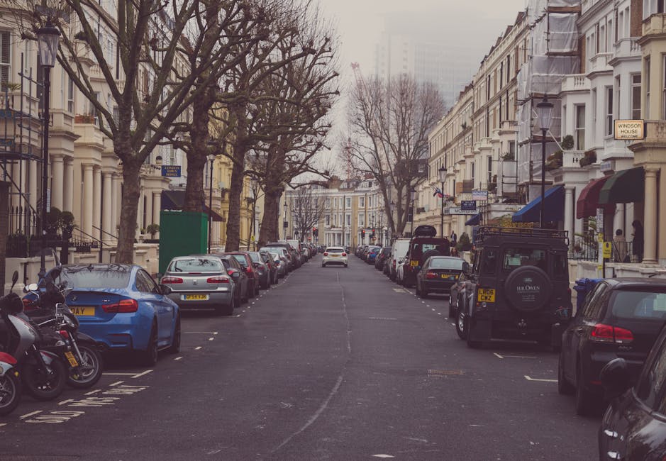 A street scene showing parked cars lining both sides of a narrow residential street with Victorian-style terraced houses featuring bay windows and decorative facades. Leafless trees are situated along the pavement, and some buildings have awnings and small front gardens, consistent with an urban home environment. The street appears to be in a city setting with overcast weather, and vehicles are parked close together, indicating limited parking space typical of narrow streets. Man With a Van Clapton’s home relocation and furniture transport services are relevant here, especially considering the tight parking conditions and street layout which require careful loading and unloading procedures, often involving moving vans or small trucks at the roadside for efficient packing and moving operations.