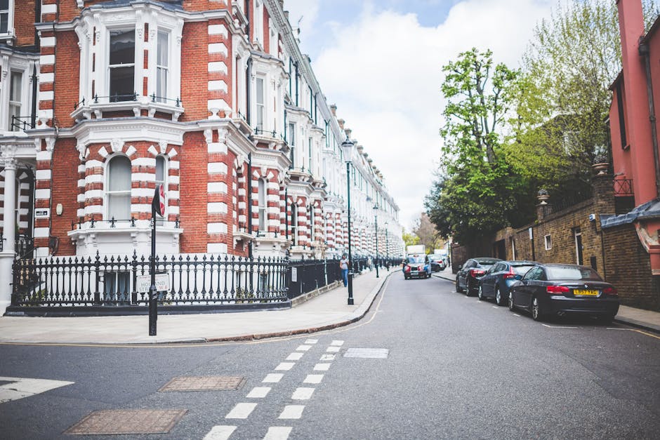 A wide street scene in a residential area featuring a Victorian-style building with red and white brickwork, decorative bay windows, and a black wrought iron fence fronting the property. The pavement is occupied by parked cars on the right side, including sedans and hatchbacks, with some vehicles partially visible or parked parallel along the curb. In the foreground, a Man With a Van Clapton crew is engaged in a home relocation process; a large cardboard box, secured with plastic wrap, is being carried by a mover who is dressed in casual clothing. The vehicle used for furniture transport—a white van—is visible further down the street, parked on the roadside with its rear doors open, ready to load or unload items. The scene is illuminated by natural daylight, with green trees lining the sidewalk on the right, and a clear blue sky overhead, emphasizing the outdoor moving and packing activity within this typical London residential setting as part of a professional removals service tailored for narrow streets and parking constraints.