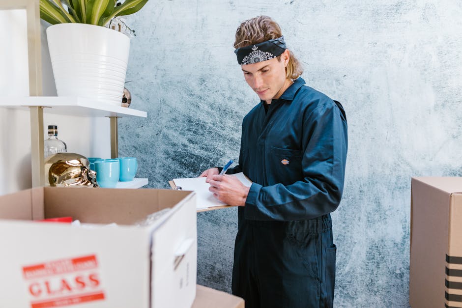 A man inside a residential property stands next to a cardboard box filled with household items, including a red-handled tool and other packing materials, as he takes notes on a clipboard. He is dressed in a dark blue work jumpsuit and wears a black bandana with a white pattern on his head. In the background, a shelving unit holds decorative objects such as a gold-colored ornament, three turquoise mugs, a glass bottle, and a white ceramic pot with a green plant. The wall behind features a textured, light-colored surface. The scene appears to be part of a home relocation or packing process, with the man from Man With a Van Clapton involved in organizing or preparing items for moving, illustrating the packing and moving stage of a house removal or furniture transport service.