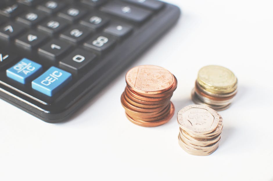 A close-up image displaying a black calculator with blue and white buttons, situated on the left side of the frame. To the right of the calculator, there are three stacks of British coins made from copper, brass, and nickel, with the coins partially overlapping each other. The coins are arranged in varying heights, with some laid flat and others leaning slightly, positioned on a plain white surface. The context suggests these coins represent expenses or costs related to house removals or moving services, as relevant to a home relocation or furniture transport process. The environment appears well-lit with natural or soft lighting, emphasizing the details of the coins and the calculator, which is typical in financial or costing discussions associated with the page about comparing removal costs at manwithavanclapton.co.uk.
