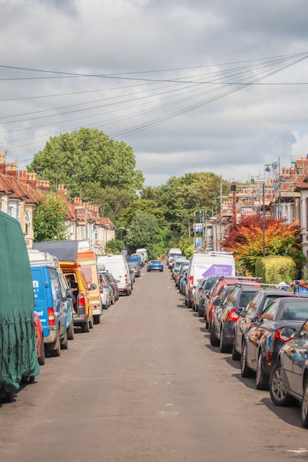 A residential street scene during daytime with closely parked cars lining both sides of the asphalt road, including small hatchbacks, sedans, and larger vans. Some vehicles are covered with protective fabric or plastic wrap, likely related to home relocation or moving preparations. The street is bordered by terraced houses with brick facades, front gardens, and small trees, under a partly cloudy sky. Overhead, multiple power lines run across the scene, and in the background, there are green, leafy trees providing a natural backdrop. This environment reflects a typical urban area where residents may experience access challenges for vehicle entry, aligned with common access problems on Upper Clapton. Such scenes are relevant to professional removals and furniture transport services provided by companies like Man With a Van Clapton, who assist with packing, loading, and transportation during home moves.