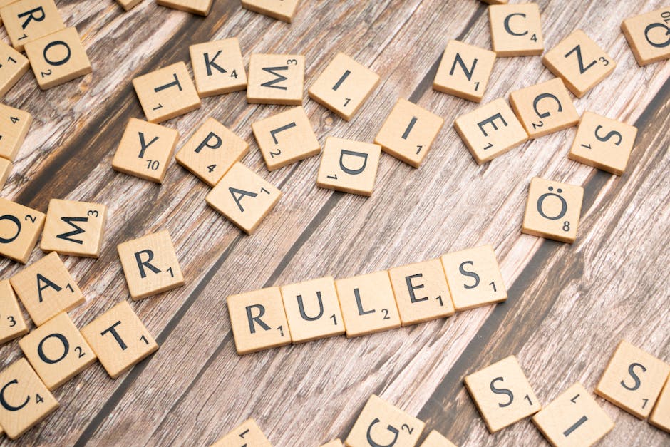 A close-up image showing numerous wooden Scrabble tiles scattered on a wooden surface, with the word 'RULES' spelled out in the center using five tiles. The tiles display black letters and numbers indicating point values, and are arranged in a slightly disorderly manner with some tiles overlapping or tilted. The setting appears to be indoors, with soft lighting illuminating the tiles and the textured wood grain beneath. This image is relevant to house removals and moving services, as it emphasizes careful planning and adherence to rules that may be important during home relocation processes. Man With a Van Clapton may utilize such imagery to subtly communicate the importance of following proper procedures when managing furniture transport and packing for a move.