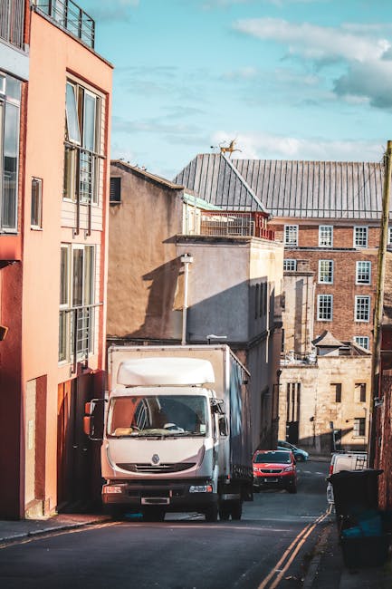 A white delivery van from Man With a Van Clapton is parked on a narrow, sloped street in an urban area during daylight, with surrounding buildings exhibiting a mix of brick and stucco facades. The van
