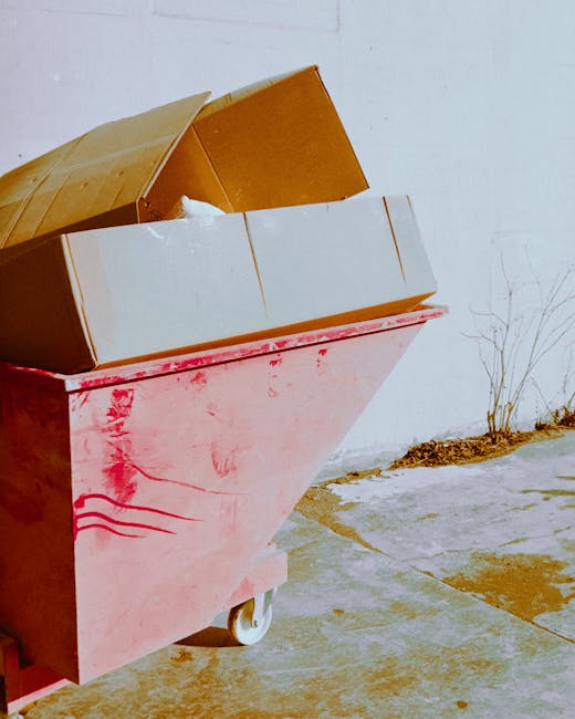 A large pink metal waste container with a slightly weathered surface, positioned on a concrete pavement outdoors. The container has a hinged lid that is open, revealing several cardboard boxes of various sizes, some with visible brown cardboard flaps, stacked inside. A piece of white packing material is partially visible within one of the boxes. The container is mounted on white wheels, and the lid is propped open to facilitate loading or unloading. To the right of the container, there is a small dried plant growing close to the wall, and the background features a plain white wall, indicating an outdoor area suitable for waste disposal or moving activities. This scene is representative of house removal preparations, with the vehicle and materials aligned with furniture transport and packing processes, as handled by Man With a Van Clapton.