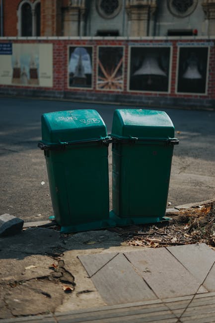 Two green wheelie bins with closed lids are positioned on the pavement outside a building, near the edge of the sidewalk. The bins are placed side by side, with a slight gap between them, and appear to be made of durable plastic material. In the background, there is a brick wall with windows framed by decorative stonework, and across the street, part of a building facade with a clock can be seen, indicating an urban environment. The pavement consists of concrete slabs with some cracks and dirt around the bins, and a small section of curb and street surface is visible. The scene is well-lit with natural daylight, suggesting daytime. The presence of the bins and the setting suggest this is a typical location for waste disposal and household rubbish collection, relevant to house removals and waste management services provided by Man With a Van Clapton, especially when considering bulky waste rules and disposal in Hackney for home relocation projects.