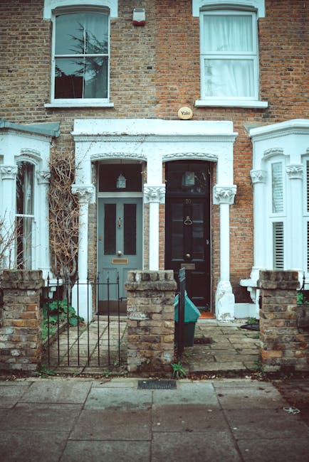 A photograph of two adjacent terraced houses showing their front entrances in Clapton. The house on the left has a grey door with a glass panel and a metal letterbox, while the house on the right features a black door with a brass door knocker and a letterbox. Both entrances are beneath white decorative porticoes with columns and moulded architraves, and there is a small front garden area enclosed by low brick walls and metal railings. The garden has paved and grassy sections, with some paving stones and a small green bag resting on the pavement in front. The upper windows have white frames, with one window open on the left. There are some bare and ivy-covered portions of the brickwork around the doors. The scene suggests a residential area suitable for home relocation or furniture transport, with the houses ready for moving services by Man With a Van Clapton.