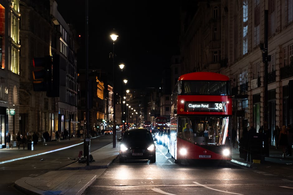 A nighttime street scene featuring a red double-decker bus with the route number 38 and destination 'Clapton Pond' displayed on the front, parked on the right side of the road alongside a black car. The street is illuminated by streetlights and the headlights of other vehicles. In the background, there are commercial buildings with lit windows and pedestrians walking along the sidewalks. The bus is part of a home relocation or furniture transport process handled by Man With a Van Clapton, highlighting typical urban logistics involved in house removals and packing and moving services, with the bus positioned near a property entrance or loading area.