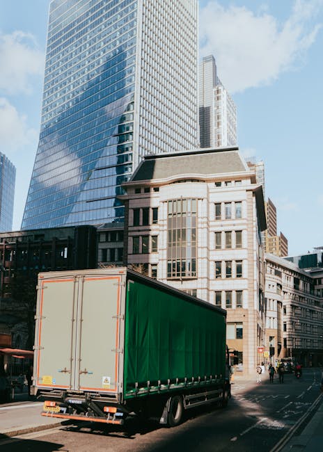 A man with glasses and a light blue shirt is seated on a wooden chair outside a white moving van, which is parked on a city street in front of a modern building with large glass windows. The van's rea
