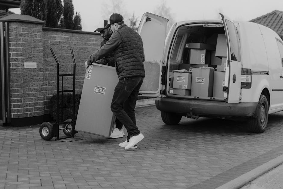 Two men are engaged in a home relocation process outside a house, preparing to load a large cardboard box onto a dolly with rubber wheels. One man is wearing a cap, glasses, and a dark quilted jacket, while the other is dressed in a jacket and white sneakers. They are lifting the box towards the open rear doors of a white delivery van parked on a paved driveway. The van's cargo area is visible, filled with several smaller cardboard boxes, some labeled with fragile stickers, and these are stacked on top of each other. The scene takes place in an outdoor setting near a brick wall and a paved pathway, with overcast skies overhead. The use of packing materials such as cardboard boxes and the equipment like the trolley and straps indicates an organized move, coordinated by [COMPANY_NAME], specializing in removals and furniture transport services, supporting seamless house relocation and moving logistics.