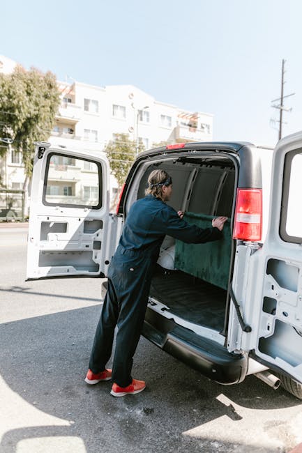 A person wearing a dark jacket, black pants, and red shoes is loading or inspecting large green equipment or furniture wrapped in protective padding inside the cargo area of a white van with open rear doors. The van is parked on a city street, with modern residential buildings and trees visible in the background, under clear weather and daylight. The scene depicts the home relocation or furniture transport process as part of a house removals service, with the individual handling items precisely inside the van, supported by the company's expertise in packing and moving logistics.