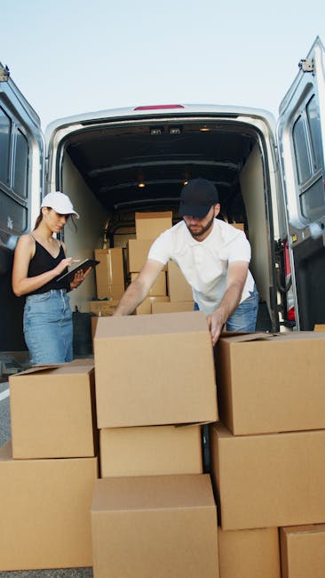 Two men are engaged in a home relocation process outside a house, preparing to load a large cardboard box onto a dolly with rubber wheels. One man is wearing a cap, glasses, and a dark quilted jacket, while the other is dressed in a jacket and white sneakers. They are lifting the box towards the open rear doors of a white delivery van parked on a paved driveway. The van's cargo area is visible, filled with several smaller cardboard boxes, some labeled with fragile stickers, and these are stacked on top of each other. The scene takes place in an outdoor setting near a brick wall and a paved pathway, with overcast skies overhead. The use of packing materials such as cardboard boxes and the equipment like the trolley and straps indicates an organized move, coordinated by [COMPANY_NAME], specializing in removals and furniture transport services, supporting seamless house relocation and moving logistics.