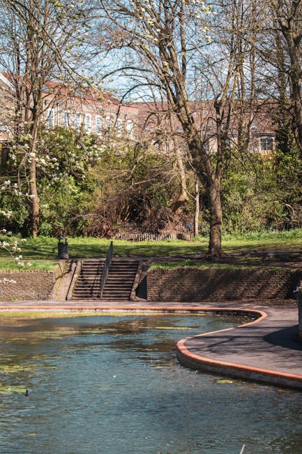 The image shows a park near a water feature with a curved brick pathway running alongside the edge of the pond or river. In the background, there are several leafless trees with bare branches, indicating early spring or late autumn, and behind the trees, there are red-brick residential buildings with multiple windows. A set of brick steps with metal railings leads from a grassy area down to the water, and the park appears well-maintained with green grass and some flowering bushes. The scene suggests a peaceful outdoor space that could be part of a house removal or home relocation process, with a focus on outdoor access and obstacle navigation, fitting within the context of local removals services provided by Man With a Van Clapton. The lighting indicates a clear day with natural sunlight illuminating the scene.
