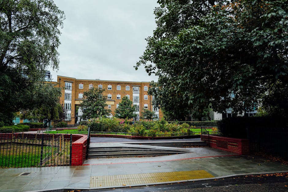 The image shows a park near a water feature with a curved brick pathway running alongside the edge of the pond or river. In the background, there are several leafless trees with bare branches, indicating early spring or late autumn, and behind the trees, there are red-brick residential buildings with multiple windows. A set of brick steps with metal railings leads from a grassy area down to the water, and the park appears well-maintained with green grass and some flowering bushes. The scene suggests a peaceful outdoor space that could be part of a house removal or home relocation process, with a focus on outdoor access and obstacle navigation, fitting within the context of local removals services provided by Man With a Van Clapton. The lighting indicates a clear day with natural sunlight illuminating the scene.