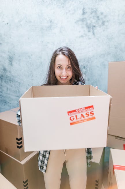 A young woman with long dark hair, smiling, holding an open cardboard box labeled 'GLASS' during a home relocation or packing process. She is standing indoors against a light blue textured wall, surrounded by several other packed cardboard boxes, some sealed with tape, in a room prepared for a house move near Chatsworth Road market Clapton. The boxes are made of sturdy brown cardboard, some with black diagonal stripes, and appear to contain household items. The woman is dressed casually in a plaid shirt and light-colored pants, and the environment is well-lit, indicating a daytime packing or loading phase, which may involve furniture transport and logistical planning in a move facilitated by Man With a Van Clapton.