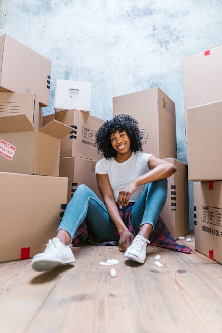 A young woman with curly black hair and a bright smile, sitting on a wooden floor surrounded by stacked cardboard moving boxes of various sizes. She is wearing a white T-shirt, light blue jeans, and white sneakers, with a plaid shirt tied around her waist. Some packing materials, such as crumpled paper, are scattered on the floor near her feet. The background features a textured light blue wall, and the scene appears to be inside a residential property during a house removal. This image illustrates the packing phase of home relocation, with the woman taking a break amid the boxes, supported by the removals services offered by Man With a Van Clapton, specializing in furniture transport and moving logistics near Chatsworth Road market.