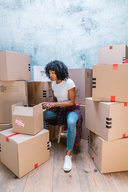 A young woman with long dark hair, smiling, holding an open cardboard box labeled 'GLASS' during a home relocation or packing process. She is standing indoors against a light blue textured wall, surrounded by several other packed cardboard boxes, some sealed with tape, in a room prepared for a house move near Chatsworth Road market Clapton. The boxes are made of sturdy brown cardboard, some with black diagonal stripes, and appear to contain household items. The woman is dressed casually in a plaid shirt and light-colored pants, and the environment is well-lit, indicating a daytime packing or loading phase, which may involve furniture transport and logistical planning in a move facilitated by Man With a Van Clapton.