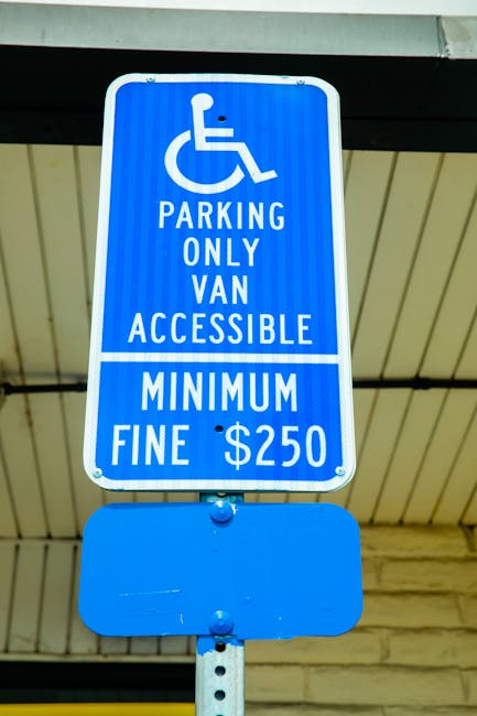 A blue parking sign mounted on a pole beneath a greenish roof in an outdoor setting, indicating that parking is reserved for vans that are accessible for disabled individuals. The sign clearly states it is for 'PARKING ONLY VAN ACCESSIBLE' with a wheelchair symbol above the text. Additional information on the sign mentions a minimum fine of $250 for violations. The background shows a roof and part of a building structure, suggesting an urban or residential area where vehicle loading zones are designated for home relocation and furniture transport. This parking restriction is relevant for relocation services, such as those offered by Man With a Van Clapton, emphasizing the importance of parking rules when conducting house removals or furniture transport in the Clapton area.