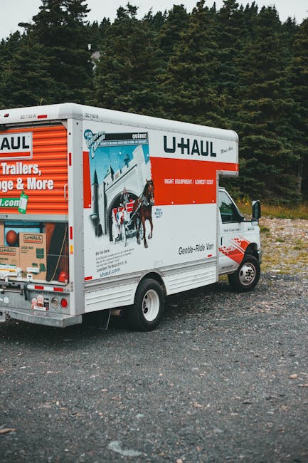 Photograph of a U-Haul moving van parked on a gravel surface near a wooded area with tall trees in the background. The van is positioned with its rear and right side visible, featuring branding and slogans related to house removals and furniture transport. The vehicle's side panel displays an illustration of a horse and carriage with a ramp, emphasizing moving services, and includes contact information. The rear of the van has a loading ramp with a partly visible box and packing materials nearby, indicating the loading process. The surroundings suggest an outdoor setting suitable for loading or unloading furniture, appliances, and packing materials such as cardboard boxes, plastic wrap, and blankets. The image supports the context of home relocation logistics managed by Man With a Van Clapton, highlighting the importance of proper vehicle and equipment use in removals services near residential areas.