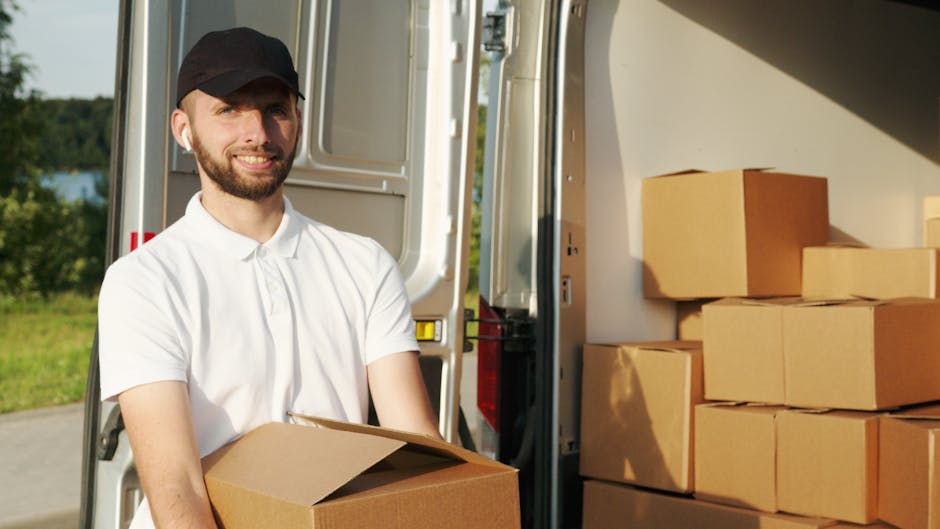 A man with a beard, wearing a black cap and a white polo shirt, is standing outside a white moving van with the rear doors open. He is holding a cardboard box and smiling at the camera. Inside the van, there are several neatly stacked cardboard boxes on the right side, some wrapped in plastic wrap, indicating packing for a home relocation. The van is parked on a paved surface near a grassy area with trees and a body of water visible in the background. The scene captures the loading process during furniture transport or packing and moving services, typical of a house removal. The lighting is natural, suggesting it is daytime, and the environment appears quiet and organized, aligning with the steps involved in professional removals by Man With a Van Clapton.
